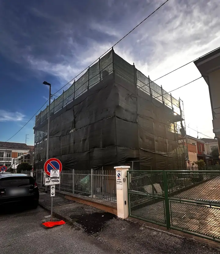 Edificio residenziale in ristrutturazione, avvolto da ponteggio e rete di protezione, sotto un cielo al tramonto